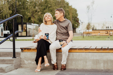 An adult mature happy couple in love sitting on bench outdoors in city street park. A blonde caucasian man and woman spend time together and drinking coffee. Senior wife and husband walking outside