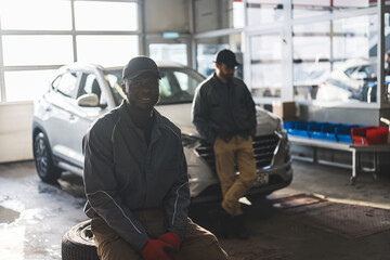 medium shot of a smiling African auto mechanic at his workshop, his co-worker leaning on the car in the background. High quality photo