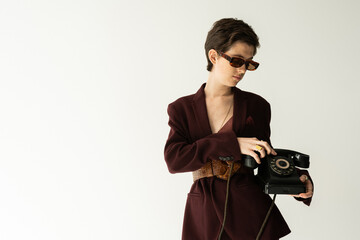 brunette woman in trendy jacket with leather belt standing with vintage phone on grey background.