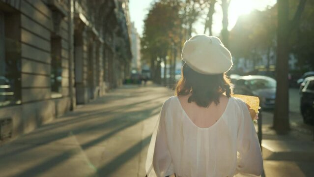 A young woman in a white beret walks with a bouquet of flowers down a sunny alley in Paris