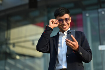 Outdoor shot of happy young businessman standing standing outdoors in urban modern city and using mobile phone