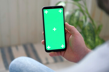 Young man sitting at home holding smartphone green mock-up screen in hand. Male person using chroma...