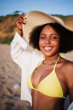 Smiling african american woman smiling with white teeth in a yellow swimsuit in a white shirt in a straw hat takes a selfie on the beach. Concept: vacation, sand, weekend, tourist tour