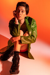 full length of dreamy woman in green leather jacket and black boots sitting and looking at camera on orange background.