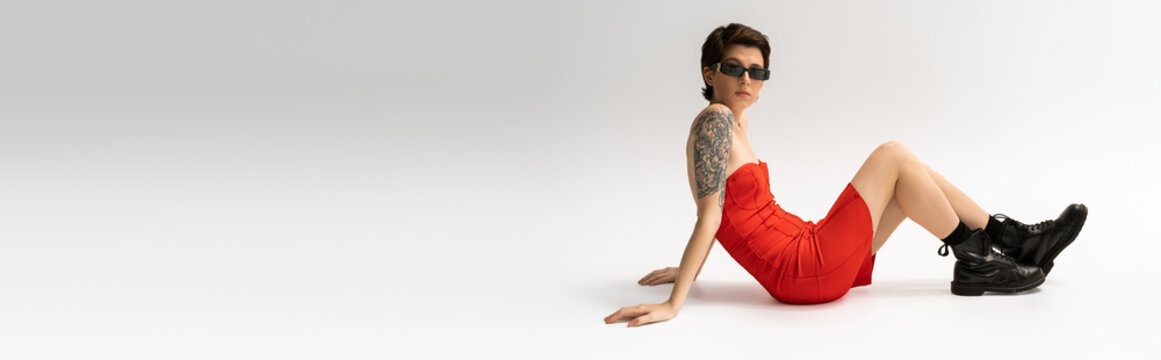full length of slim tattooed woman in black boots and red corset dress sitting on grey background, banner.