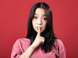 Naklejka premium A portrait of a young Indonesian (Asian) woman wearing a pink shirt, looking like she's telling someone to be quiet. Isolated with a red background