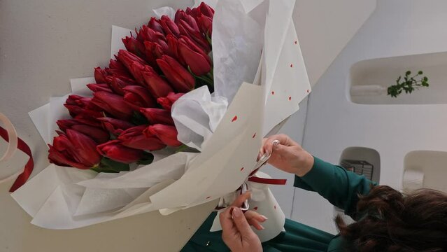 Caucasian lady florist tying a ribbon at the base of bouquet of tulips. A professional makes a fresh bouquet. Small business. Vertical video.