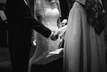 Grayscale closeup of bride and bridegroom holding each other's hand and standing in front of priest