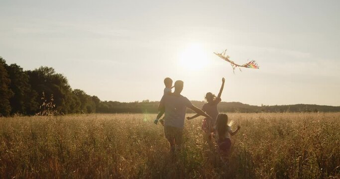 Happy young active family of four have fun in a golden wheat field at sunset. Mom, dad, son and daughter run together holding colorful kite during golden hour.