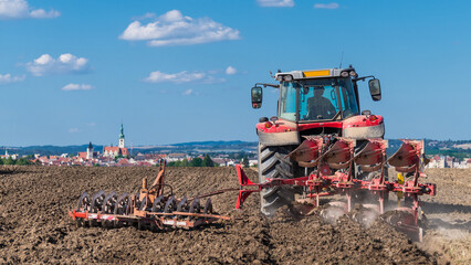 Obraz premium Red tractor with plow and disc harrow in brown arable soil under summer blue sky. Farm vehicle at working on dusty field with skyline of hussites Tabor city and church tower in scenic Czech landscape.