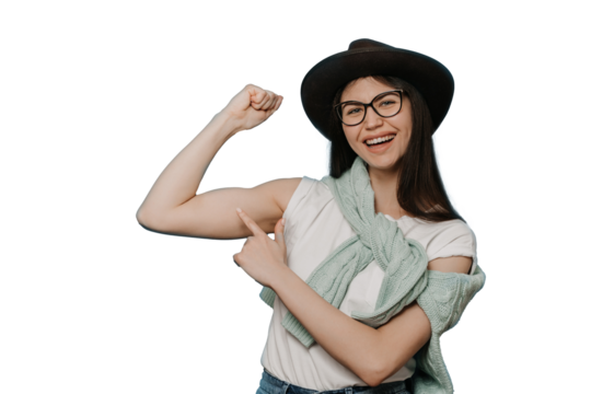 Adorable young brunette girl dressed in white t-shirt, brown hat and knitted sweater tied around her neck, shows her biceps and points at it by another hand with wide smile over transparent background