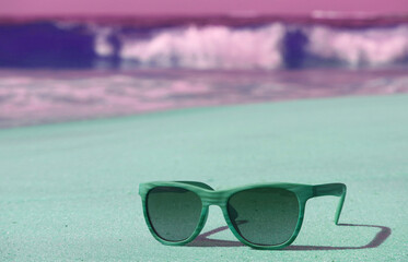 Fototapeta na wymiar Pop art styled green and purple sunglasses on the sandy beach with splashing waves in background