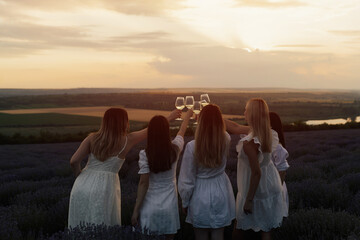 Group of four young girlfriends enjoying a picnic standing outdoors on a lavender field and clinking glasses of  wine at sunset.