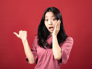 A studio portrait of a young Indonesian (Asian) woman wearing a pink shirt and pointing with her thumb. Isolated with a red background.