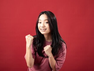 A studio portrait of a young Indonesian woman (Asian) wearing a pink shirt, seen celebrating and raising both hands. Isolated with a red background