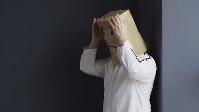 A sad man in a white shirt with a bag on his head, with a drawn crying emoticon, stands near the wall, holds his head and cries. Emotions and gestures.