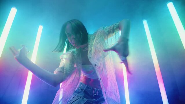 Stylish slim woman dance with hands backlit colorful neon light lamps, beautiful female in trendy outfit dancing in night club, creative performer, haze and glowing tube lights, slow motion wide shot
