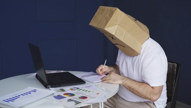 A man - a businessman in a white shirt with a paper bag on his head, with a sad smiley face drawn, works at a computer with documents, makes notes in a notebook. Emotions and gestures.