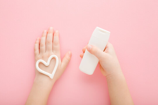 White heart shape on baby girl hand. Light pink table background. Pastel color. Hand holding little tube of moisturizing cream. Closeup. Point of view shot. Care about child hands skin. Top down view.