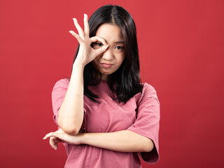 A studio portrait of a young Indonesian (Asian) woman wearing a pink shirt and making the OK sign with her hand in front of her face. Isolated with a red background