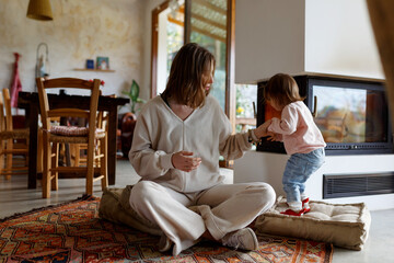 Happy young mother and toddler daughter spending time together on floor near fireplace at home. Cozy interior