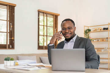 Fototapeta na wymiar Portrait of handsome African black young business man working on laptop and talking phone at office desk.