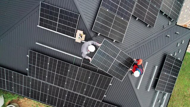 Technicians building solar panel system on roof of house. Drone view of men workers in helmets carrying photovoltaic solar module outdoors. Concept of alternative and renewable energy.