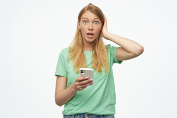 Naklejka premium Indoor studio portrait of young ginger female with freckles confused, looking at phone, touching her head isolated over background