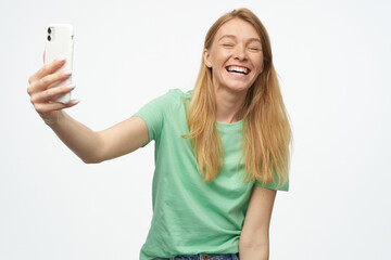 Indoor studio portrait of young ginger female with freckles making selfie and smiles broadly isolated over background