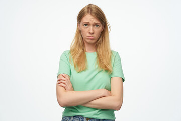 Fototapeta premium Indoor studio portrait of young ginger female with freckles looking into camera, keeps hands folded with sad facial expression, isolated over background