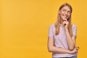 Indoor studio portrait of young ginger female with freckles thoughtfully looking aside, smiles broadly, isolated over yellow orange background