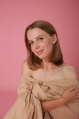 beautiful sexy blonde girl on pink background studio shot. A girl wrapped in a beige cloth sits on a pink background. beautiful girl model with make-up, stylish shooting