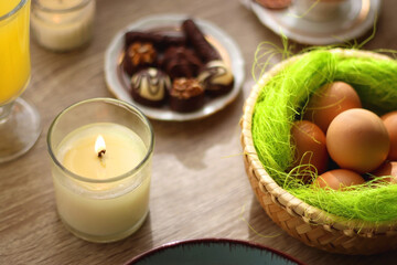 Easter eggs in the basket, bowl of cookies, chocolate pralines, Easter bunny figurine, cups of tea, glasses of juice, flowers and lit candles on the table. Selective focus.