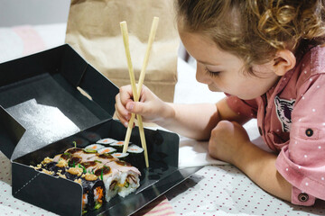 A cute white Caucasian little girl picking up some sushi maki for lunch with chopsticks from a takeaway box on the kitchen table at home