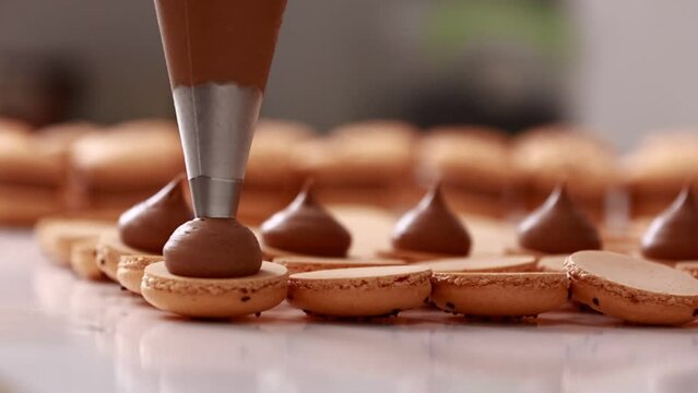 Making french desert macarons. Chef hands with confectionery bag add cream to the ready halves of brown chocolate macaroons at pastry shop. cooking, food and baking concept.