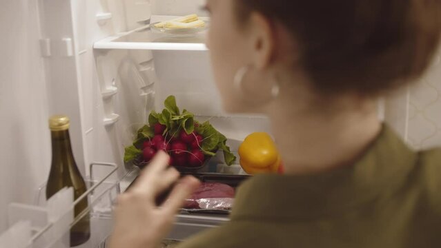 Back view of young Caucasian woman putting food products in fridge while cooking at home