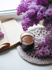 Obraz na płótnie Canvas Morning coffee and a book on the windowsill. Blooming spring lilac in a vase. Knitted napkin
