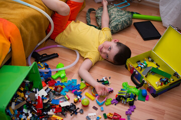 A cute six year old child boy lies on the floor in a pile of his toys. A mess in the children's room.