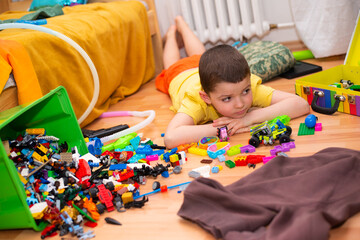 A sad six-year-old boy lies on the floor in the mess of his room in a pile of toys