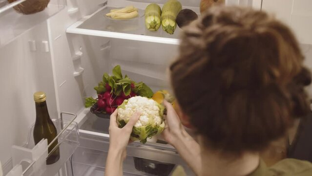 Over the shoulder shot of young Caucasian woman putting fresh groceries on shelf in fridge at home