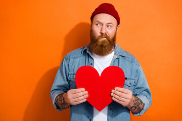 Photo of ponder young man wear beanie hat hold red paper love symbol postcard look mockup valentine day gift isolated on orange color background