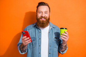 Photo of hipster man red beard hair coffee break hold plastic cup drinking relax time chilling browsing phone isolated on orange color background