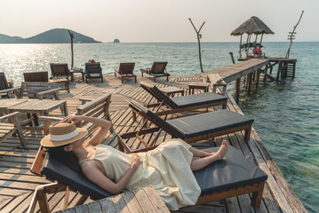 Woman traveler wear white dress and relax on wooden beach chair in summer holiday at hidden bar on the sea