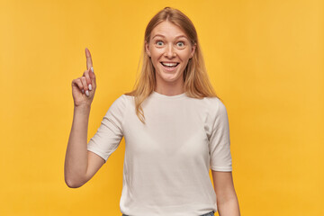 Indoor studio portrait of young ginger female with freckles points with a finger upwards with surprised facial expression, isolated over yellow orange background