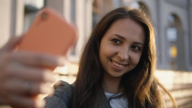 Woman takes selfies using smartphone camera outdoors. Girl wearing gray jacket and wireless earphones smiles while taking photos with her phone it the street at sunset 