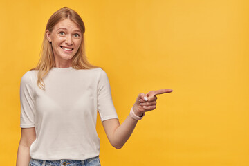 Fototapeta premium Indoor studio portrait of young ginger female with freckles points with a finger aside, isolated over yellow orange background