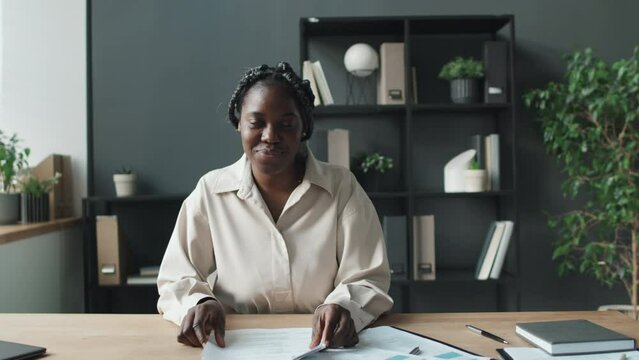 Medium shot of African American HR finishing looking through CVs, sitting in light office beside window, then looking at camera, broadly smiling, front view