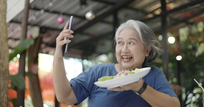 An elderly Asian woman is happily using a smartphone selfie camera to take a picture of food in the garden of her tiny house. Internet communication technology to connect with family