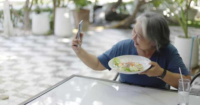 An elderly Asian woman is happily using a smartphone selfie camera to take a picture of food in the garden of her tiny house. Internet communication technology to connect with family