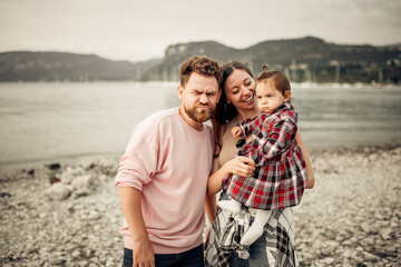 Young happy family spending their vacation on Italian Garda lake coast, bearded daddy making unpleased faces, smiling mom, their little toddler girl in red checkered dress pointing with finger